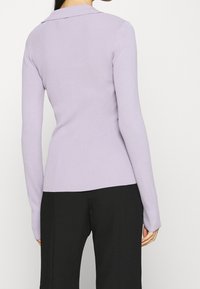 Fialový žebrovaný top s dlouhým rukávem a límečkem, přiléhavý střih a texturovaná látka, spárovaný s černými kalhotami. Minimalistický vzhled.
