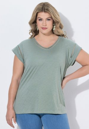 Kobieta o falowanych blond włosach, ubrana w jasnozielony krótki rękaw z wycięciami i niebieskie dżinsy, stoi z jedną ręką na biodrze.