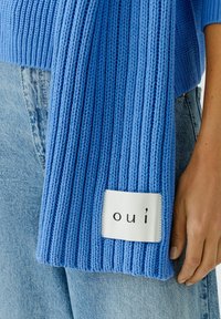 Blauwe geribbelde gebreide trui met een textuurafwerking, voorzien van een wit label met de tekst "ou'i." De trui is gedeeltelijk in lichtblauwe spijkerbroek gestoken.