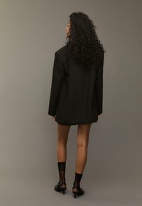 Kvinde med langt krøllet hår iført en oversized sort blazer, gennemsigtige sorte ankelstrømper og sorte hæl sandaler, stående mod en ensfarvet baggrund.