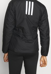 Vue arrière d'une personne portant une veste noire avec trois rayures verticales blanches sur le haut du dos et un legging noir.