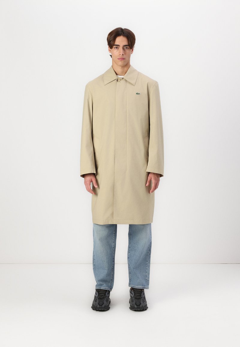Beige, lang regnfrakke med krave, frontlukkede knapper og sidelommer. Båret over blå jeans og sorte sneakers med et lille logo.