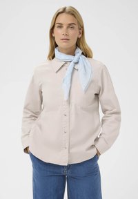 Camisa de botón color beige claro con cuello, un bolsillo en el pecho y mangas largas. Llevada con una bufanda atada de color azul claro. Jeans azules completan el look.