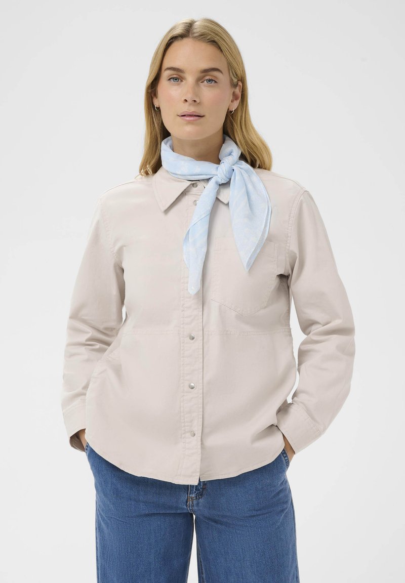 Camisa de botón color beige claro con cuello, un bolsillo en el pecho y mangas largas. Llevada con una bufanda atada de color azul claro. Jeans azules completan el look.