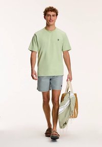 Heleroheline lühikeste varrukatega särk väikese logoga, ühendatud triibuliste lühikeste pükste ja sandaalidega, käes kootud kott koos rätikuga.