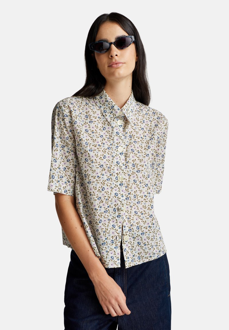 Vrouw die kattensoorten zonnebrillen draagt en een kortarmige overhemd met een klein bloemenpatroon, gecombineerd met donkerblauwe jeans.
