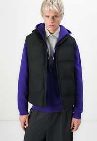 Czarna kamizelka puchowa z wysokim kołnierzem, noszona na fioletowym hoodie i koszulce w paski. Posiada zamek błyskawiczny oraz luźny krój.