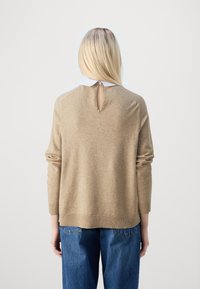 Blondýna v béžovém svetru s výstřihem ve tvaru klíče a bílým límečkem, v volných modrých džínách, stojící zády k bílé pozadí.