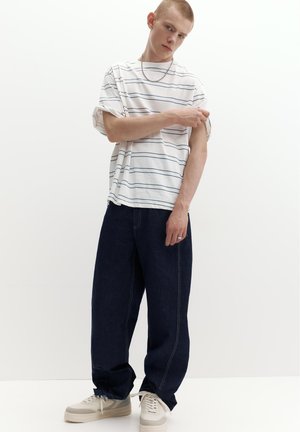 Junger Mann in weißem gestreiftem T-Shirt, der seinen Ärmel hochkrempelt, trägt dunkle lockere Jeans und weiße Sneakers, steht vor einem weißen Hintergrund.