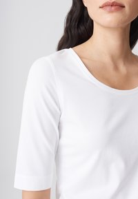 Femme portant un t-shirt blanc uni à manches courtes, montrant le décolleté, l'épaule et une partie du visage sur un fond gris clair.