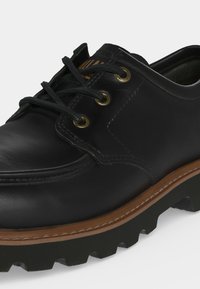 Chaussure noire en cuir à lacets avec semelle cousue marron et semelle extérieure épaisse crantée, vue en gros plan sur fond blanc.