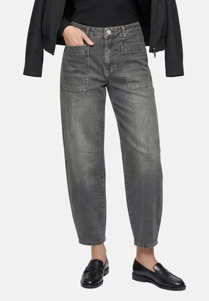 Frau trägt hoch taillierte, ausgewaschene graue Jeans mit übergroßen Vordertaschen, schwarze Loafer und eine schwarze Jacke, die über die Schultern gelegt ist.