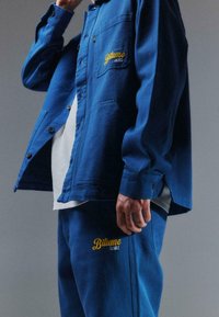 Chemise bleue à boutons avec une poche poitrine et un logo brodé jaune, associée à un pantalon bleu assorti portant la même marque.