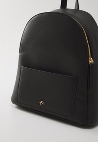 Sac à dos en similicuir noir avec poche avant, fermeture éclair dorée et petit logo doré sur la poche, présenté légèrement incliné sur fond blanc.