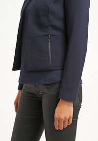 Veste cintrée bleu marine avec une poche zippée à l'avant, manches longues et une texture de tissu lisse. Portée avec un pantalon noir.