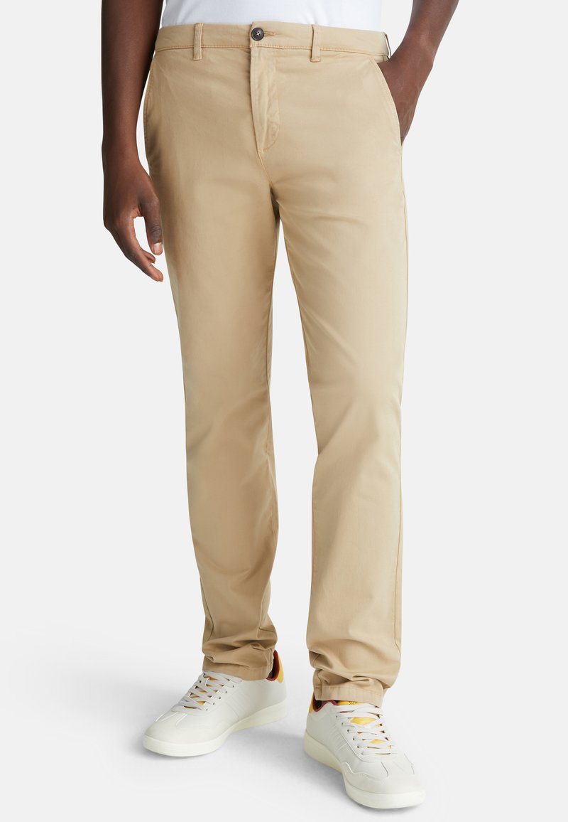 Chino in cotone beige con davanti piatto, due tasche laterali e chiusura con bottone, abbinati a sneaker bianche con accenti multicolori.