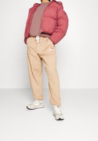 Fuzzy vinröd jacka över en grå sweatshirt, tillsammans med beige joggingsbyxor med en logotyp, och vita sneakers med grå detaljer.