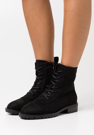 Une paire de bottines noires en daim à lacets, avec fermeture éclair sur le côté et talon bas, portées sur des jambes nues sur fond blanc.