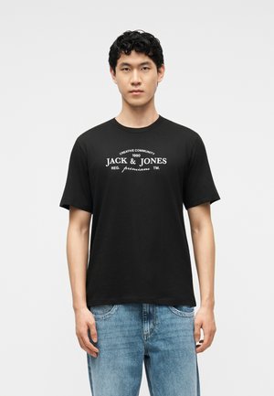 Νεαρός άνδρας με κοντά σγουρά μαύρα μαλλιά, φορώντας μαύρο μπλουζάκι "Jack & Jones" και μπλε τζιν, στέκεται μπροστά σε ένα απλό φωτεινό φόντο.