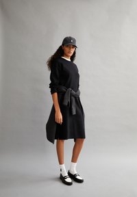 Abito felpa nero con colletto bianco, maglione grigio annodato in vita, calze bianche e scarpe nere con accenti bianchi.