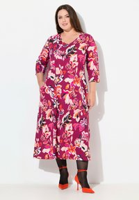 Vestido maxi en color púrpura con patrones florales en rosa, naranja y negro; presenta mangas de tres cuartos y bolsillos laterales. Combinado con tacones puntiagudos rojos.