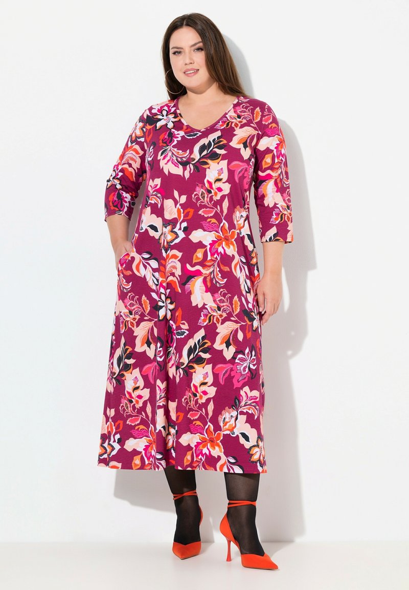 Vestido maxi en color púrpura con patrones florales en rosa, naranja y negro; presenta mangas de tres cuartos y bolsillos laterales. Combinado con tacones puntiagudos rojos.