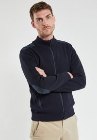 Pull en zip bleu marine, avec un col montant, des bords côtes aux poignets et des renforts aux coudes dans une teinte contrastante. Texturé en tricot avec une coupe ajustée.