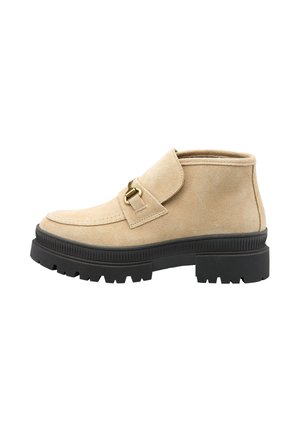 Bottine en daim beige avec un rabat, accents en métal doré et semelle en caoutchouc noir texturé présentant des rainures profondes pour une meilleure adhérence.
