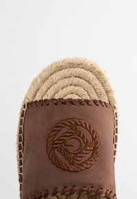 Brown leather slip-on sandal with a woven jute sole. Features embroidered circular design and decorative stitching along the edge.