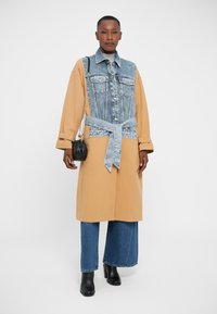 Denim i bež oversized kaput s dugim rukavima, remenom oko struka, prednjim džepovima i ležernim krojem. Uparen sa širokim trapericama i crnim čizmama.