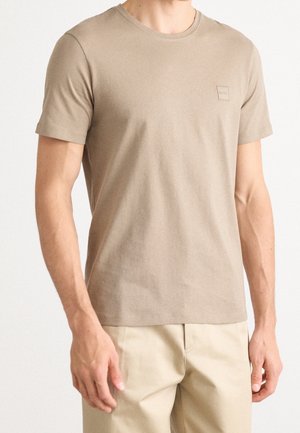 Homme portant un t-shirt beige ajusté à manches courtes et un pantalon beige clair, debout devant un fond blanc uni, les mains relâchées.