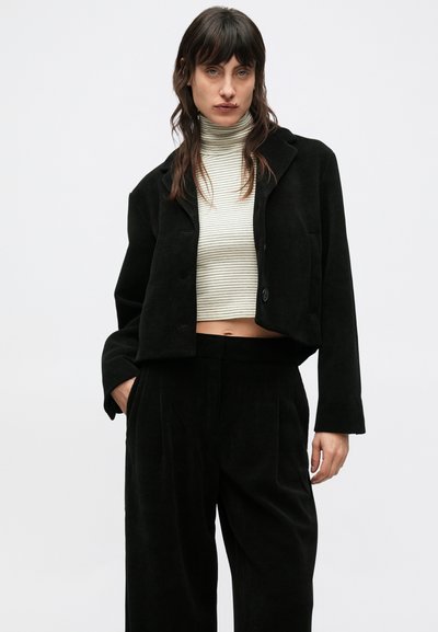 Fekete kordbársony rövidített dzseki gombos záródással, csíkos garbóval párosítva. A modell egy egyszínű fehér háttér előtt áll.