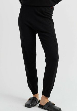 Personne portant un pantalon de jogging noir ajusté et des chaussures noires sans lacets avec un détail de boucle dorée, debout sur un fond clair uni.