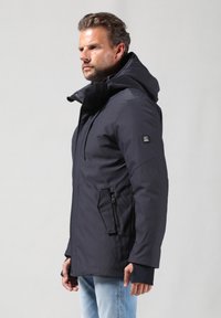 Marineblå, isoleret jakke med hætte, der har en lynlåslomme på siden, ribbede manchetter og et strømlinet, moderne design.