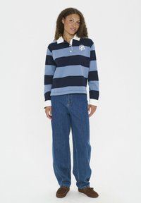 Sininen ja tummansininen raidallinen pitkähihaisen polo, jossa on valkoinen kaulus, yhdistetty löysiin sinisiin farkkuihin ja ruskeisiin slip-on-kengiin. Hienovarainen logo rinnassa.