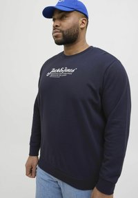 Marineblaues Sweatshirt mit langen Ärmeln, gerippten Bündchen und Rundhalsausschnitt; verfügt über weißen Logo-Text auf der Brust; kombiniert mit hellblauen Jeans.