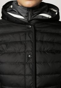 Sort pufferjakke med lynlåse foran og trykknaplukninger, med et struktureret, quiltet design og en foret hætte, der afslører grå detaljer.