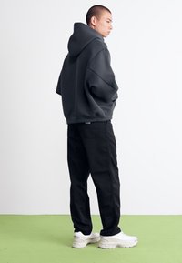 Ung mand står sidelæns iført en sort oversized hoodie, sorte bukser og hvide sneakers på et grønt gulv.