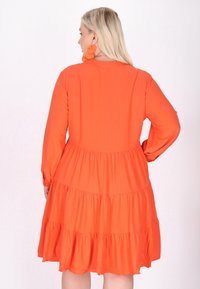 Langärmliges orange Kleid mit einem gestuften Design, das eine fließende Stofftextur und Raffungen an der Taille und am Saum aufweist.