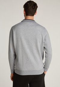 Hombre vestido con un suéter de manga larga en gris claro, con puños y dobladillo acanalados, de pie con las manos en los bolsillos, visto desde atrás.