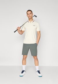 Πολο μπλούζα σε κρέμα με κόκκινες λεπτομέρειες, συνδυασμένη με γκρι σορτς. Το μοντέλο κρατάει μια γκολφ κλαμπ, ντυμένο με λευκά παπούτσια και κάλτσες με μπλε λεπτομέρειες.