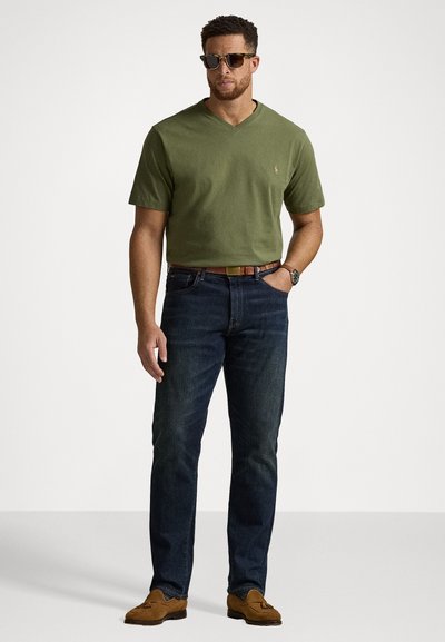 Zielona oliwkowa koszulka z krótkim rękawem i dekoltem w serek, niebieskie dżinsy, brązowy pasek i brązowe mokasyny; mężczyzna noszący okulary przeciwsłoneczne, stojący na jednolitym tle.