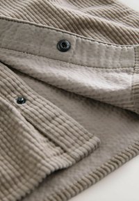 Jasnobrązowa, teksturowana tkanina charakteryzująca się poziomymi prążkami, czarnymi guzikami i gładkim wykończeniem, prezentująca button-up z finezyjnymi detalami.