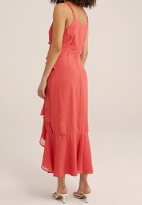 Coral dress made of lightweight fabric, featuring ruffles along the side, a flared hem, and adjustable spaghetti straps.