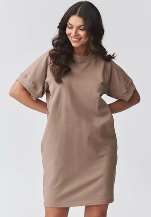 Uśmiechnięta kobieta o długich, ciemnych, falowanych włosach, ubrana w luźną, beżową sukienkę z krótkim rękawem, stojąca z rękami za plecami na jednolitym tle.