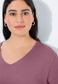 V-hals, kortmouw top in gedempt mauve stof. Soepele textuur met subtiele glans, heeft gestikte randen. Het model draagt zilveren creolen.