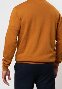 Homme portant un pull orange brûlé à manches longues et un pantalon bleu marine foncé, vu de dos sur un fond clair uni.
