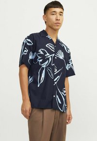 Camicia navy blu a maniche corte con motivi floreali bianchi e azzurri, con chiusura a bottoni sul davanti e silhouette rilassata. Tessuto setoso e leggero.