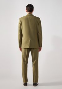 Completo verde oliva, vestibilità sartoriale, giacca monopetto con rever a scossa, pantaloni abbinati, tessuto liscio, con scarpe con accenti scuri.