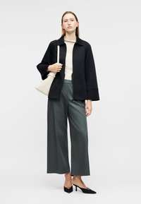 Γυναίκα με μαύρο σακάκι, κρεμ τοπ, φαρδύ γκρι παντελόνι, μαύρα τακούνια, κρατώντας λευκή τσάντα ώμου μπροστά σε γκρι φόντο.